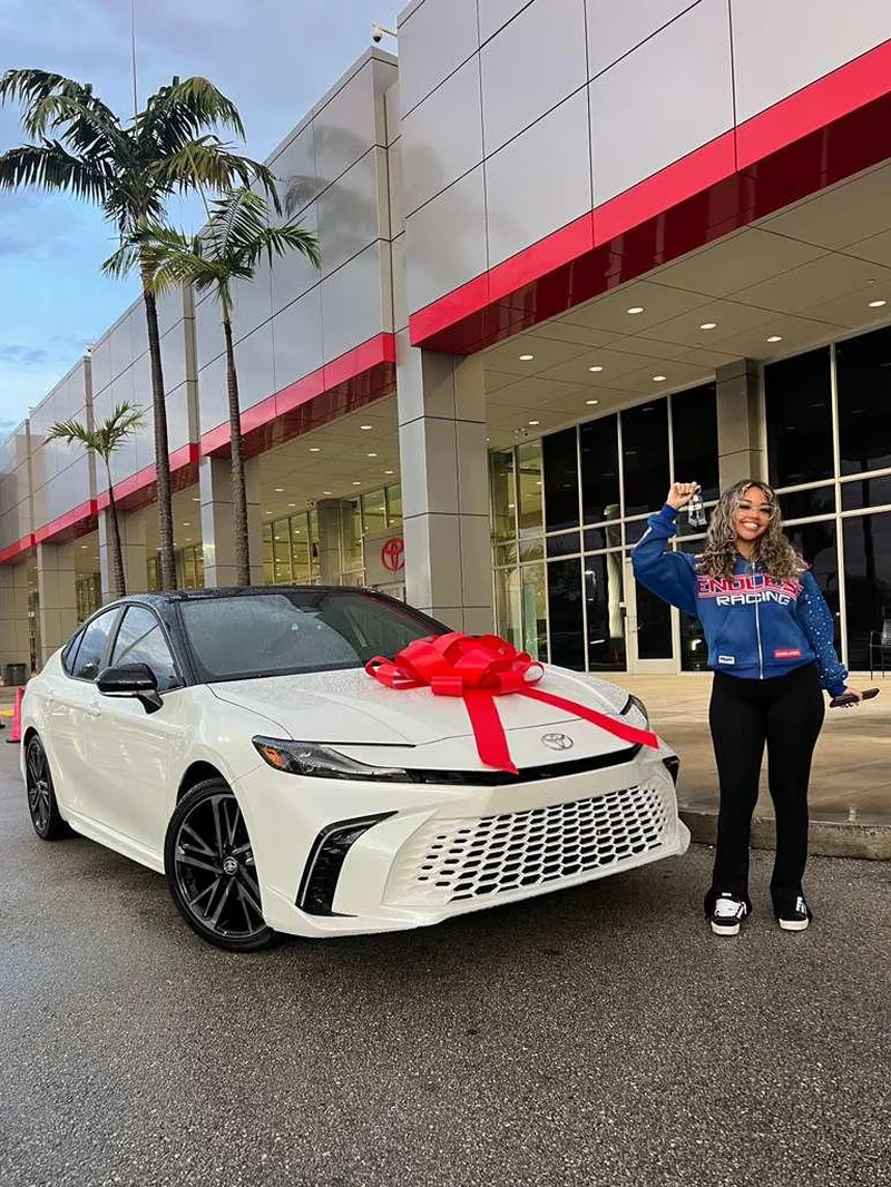 A customer holding up her keys next to a new white Toyota Camry with a red delivery bow