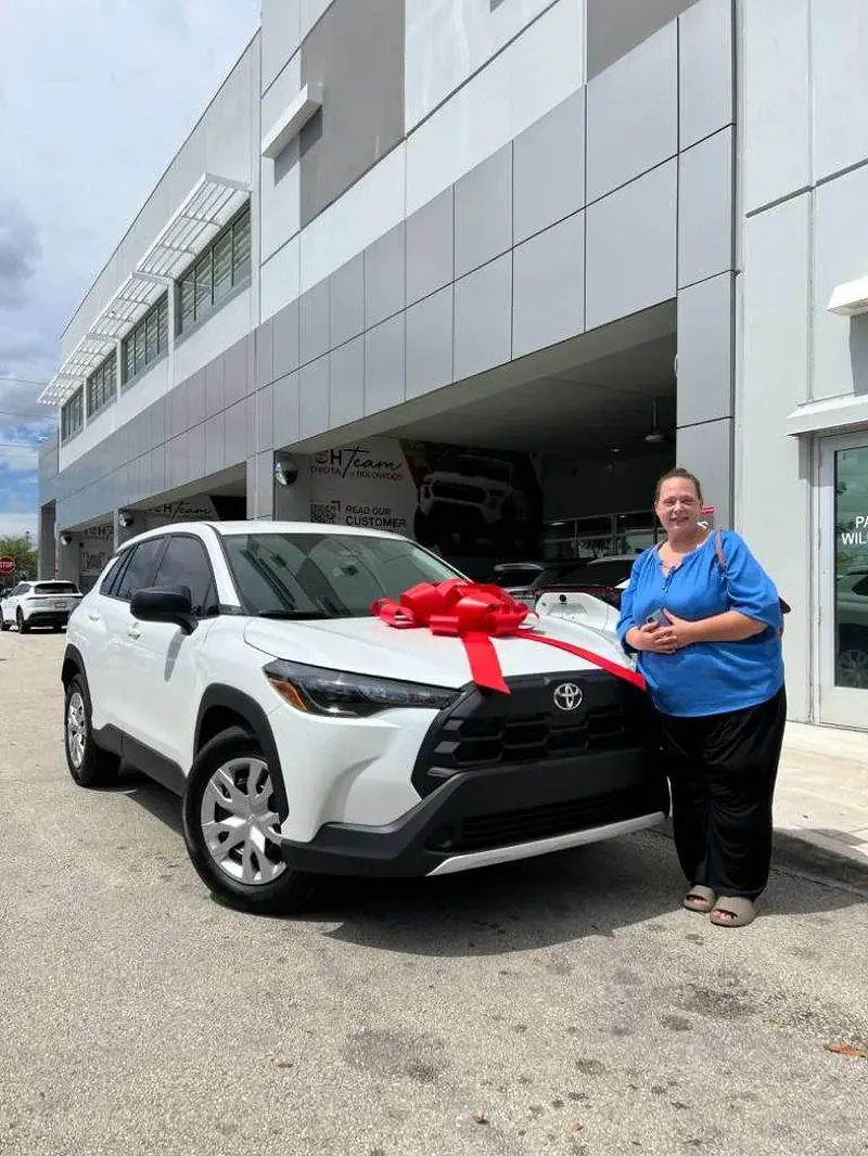 A customer smiling beside her new white Toyota Corolla Cross with a red delivery bow